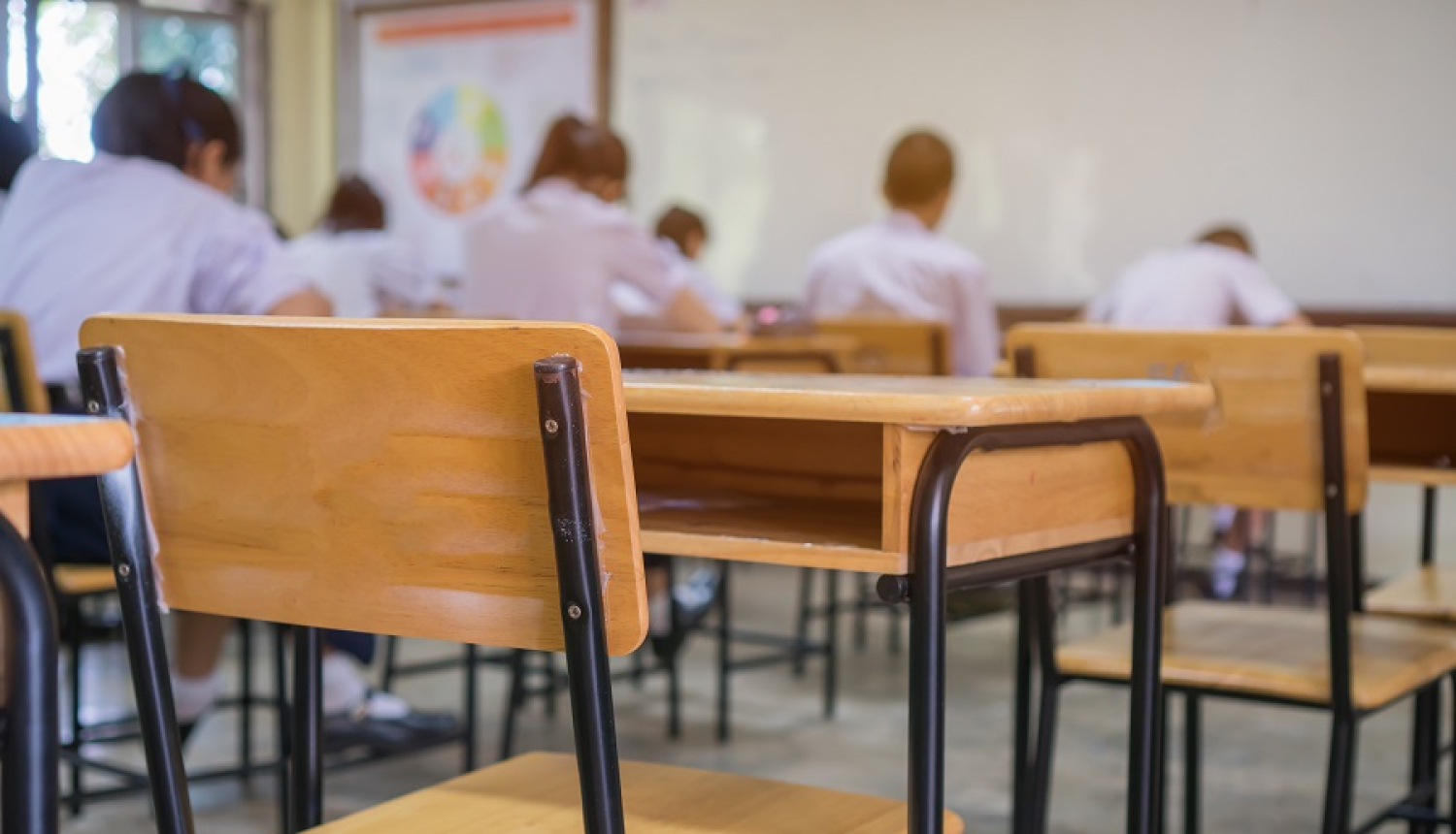 Skolas klase, skolas galdi un krēsli. Fonā ar mugurām sēdoši skolēni 