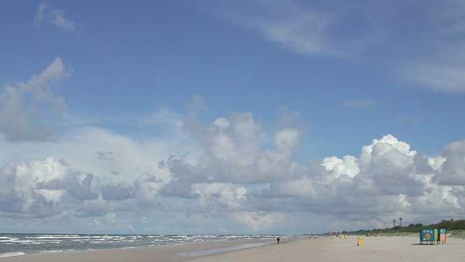 Pludmale, zilas debesis un jūra. Fonā pastaigājas daži cilvēki