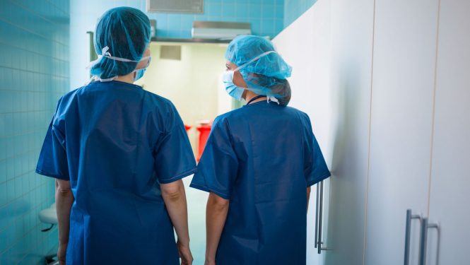 Ar muguru stāvoši divi medicīnas darbinieki zilā darba apģērbā