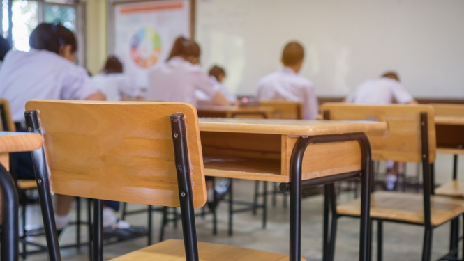 Skolas klase, skolas galdi un krēsli. Fonā ar mugurām sēdoši skolēni 