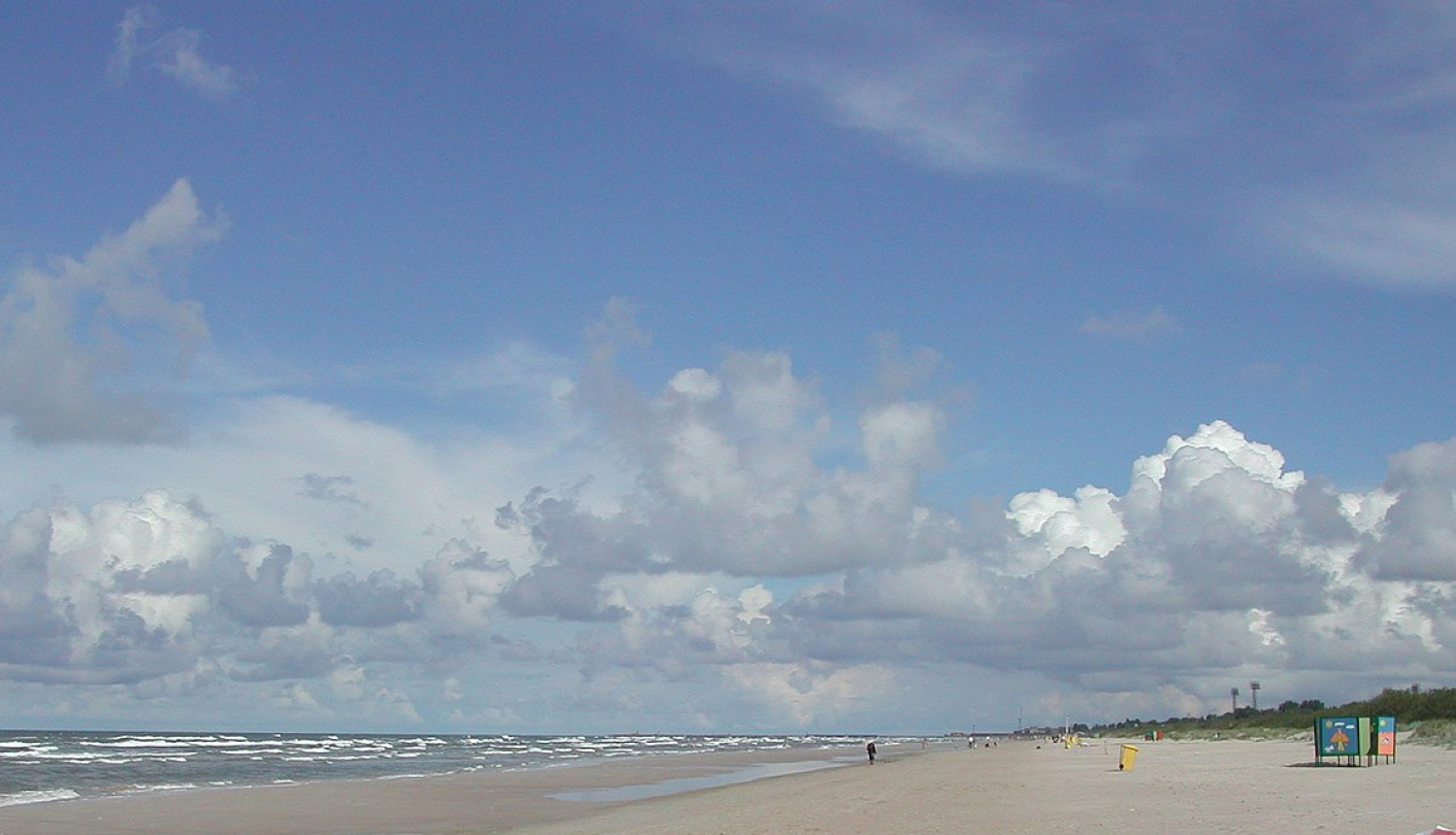 Pludmale, zilas debesis un jūra. Fonā pastaigājas daži cilvēki