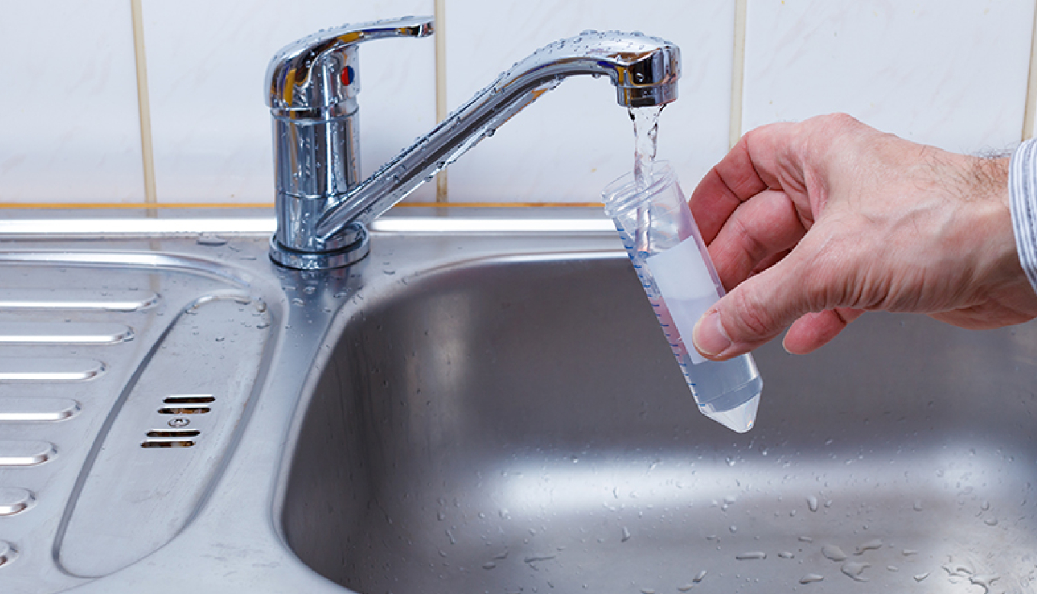 Tekošs ūdens krāns, cilvēka roka tur laboratorijas stobru zem krāna 